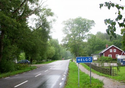 En väg med grönska, en blå skylt där det står Helgö.